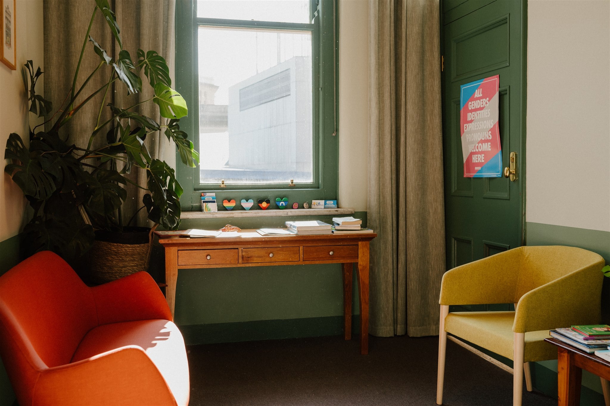 Cushioned chairs to wait in next to a large, bright window. Plants and an inclusive poster and flags decorate the space.